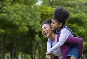 African American mother is playing piggyback riding with her young daughter while having a summer picnic in the public park